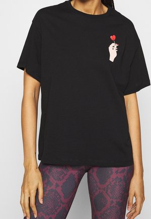 Kvinde iført en sort t-shirt med en lille grafik af en hånd, der holder en rød hjerteformet slikkepind på brystet, kombineret med lilla mønstrede leggings.