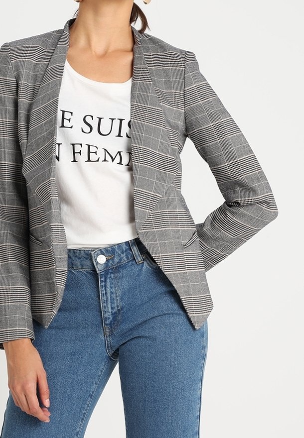 Femme portant un blazer gris à carreaux sur une chemise blanche avec un texte noir partiel, assorti à un jean bleu taille haute.