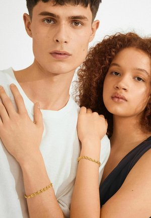 Young man and woman standing close, both wearing gold chain bracelets, the man in a white shirt and the woman in a black top.