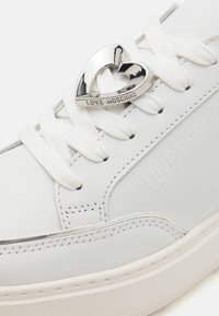 Baskets blanches avec une tige en cuir lisse, contrastées par un emblème en forme de cœur argenté brillant et des lacets blancs texturés. Marque estampillée visible.
