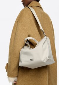 Beżowa torba o gładkiej, teksturowanej powierzchni i krótkich uchwytach, noszona na brązowym płaszczu. Na przodzie znajduje się czarne logo marki.