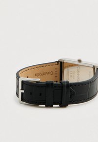 Bracelet de montre en cuir noir avec une texture lisse embossée croco, doté d'une boucle en métal argenté et d'un boîtier carré en acier inoxydable.