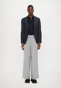 Navy-blaues, gestreiftes Blazer, dunkles Hemd und hellgraue, weit geschnittene Hose, ergänzt durch schwarze Schuhe, die eine strukturierte, moderne Silhouette schaffen.