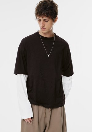 Zwarte T-shirt met korte mouwen, gedragen over een witte overhemd met lange mouwen, in combinatie met beige broeken. Heeft een eenvoudig ontwerp en een relaxte pasvorm.