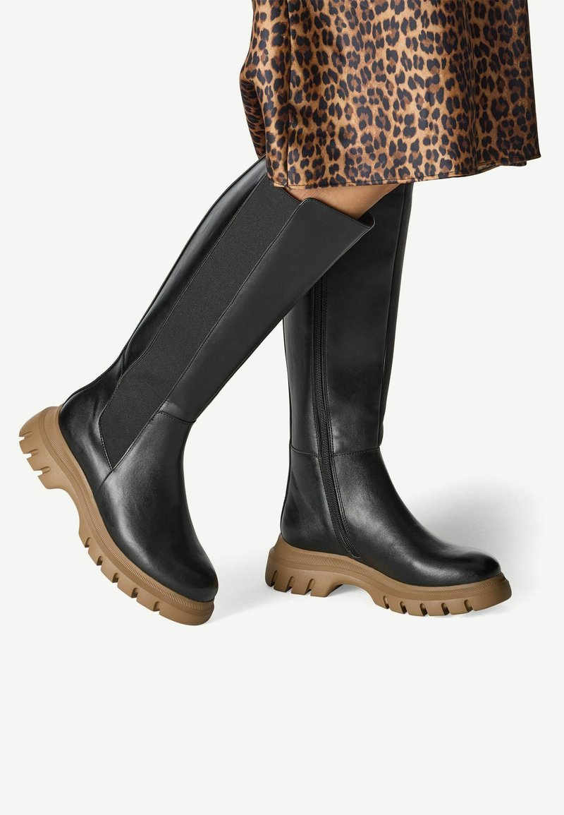 Bottes noires en cuir jusqu'au genou avec une semelle en caoutchouc épaisse de couleur tan et des panneaux élastiques sur les côtés pour un ajustement parfait ; texture lisse et design épuré.