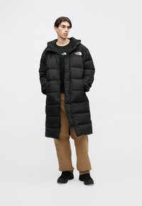 Crna puhasta jakna s kapuljačom, s prednjim patent-zatvaračem, podijeljena na nekoliko dijelova i s ukrasnim logotipima. Nošena preko crne majice i smeđih hlača.