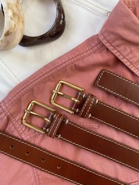 Bruna läderbälten med guldfärgade spännen på en rosa tygbakgrund; inkluderar två cirkulära resinringar i ljusa och mörka färger.