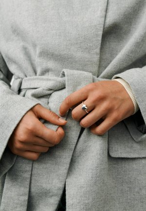 Ruky, ktoré viažu sivý opasok kabáta, so strieborným prsteňom v tvare srdca na prostredníku pravej ruky, viditeľný béžový manžetový rukáv svetra pod ním.