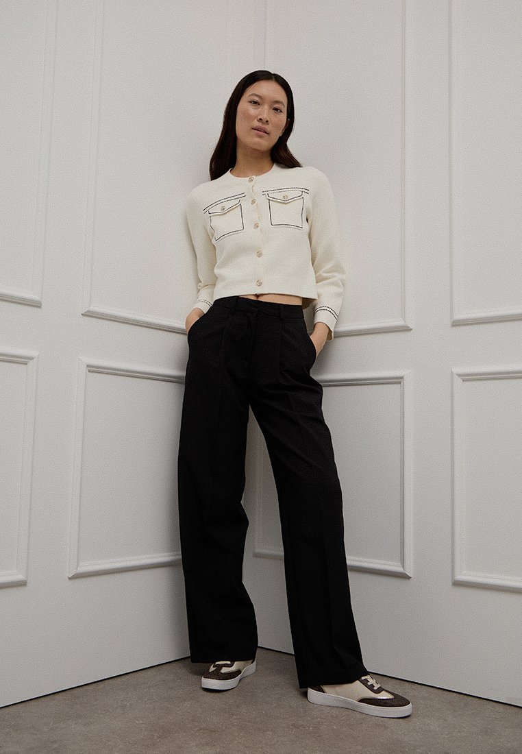 Cardigan court crème avec deux poches avant et un passepoil noir, associé à un pantalon noir taille haute plissé et des baskets décontractées.