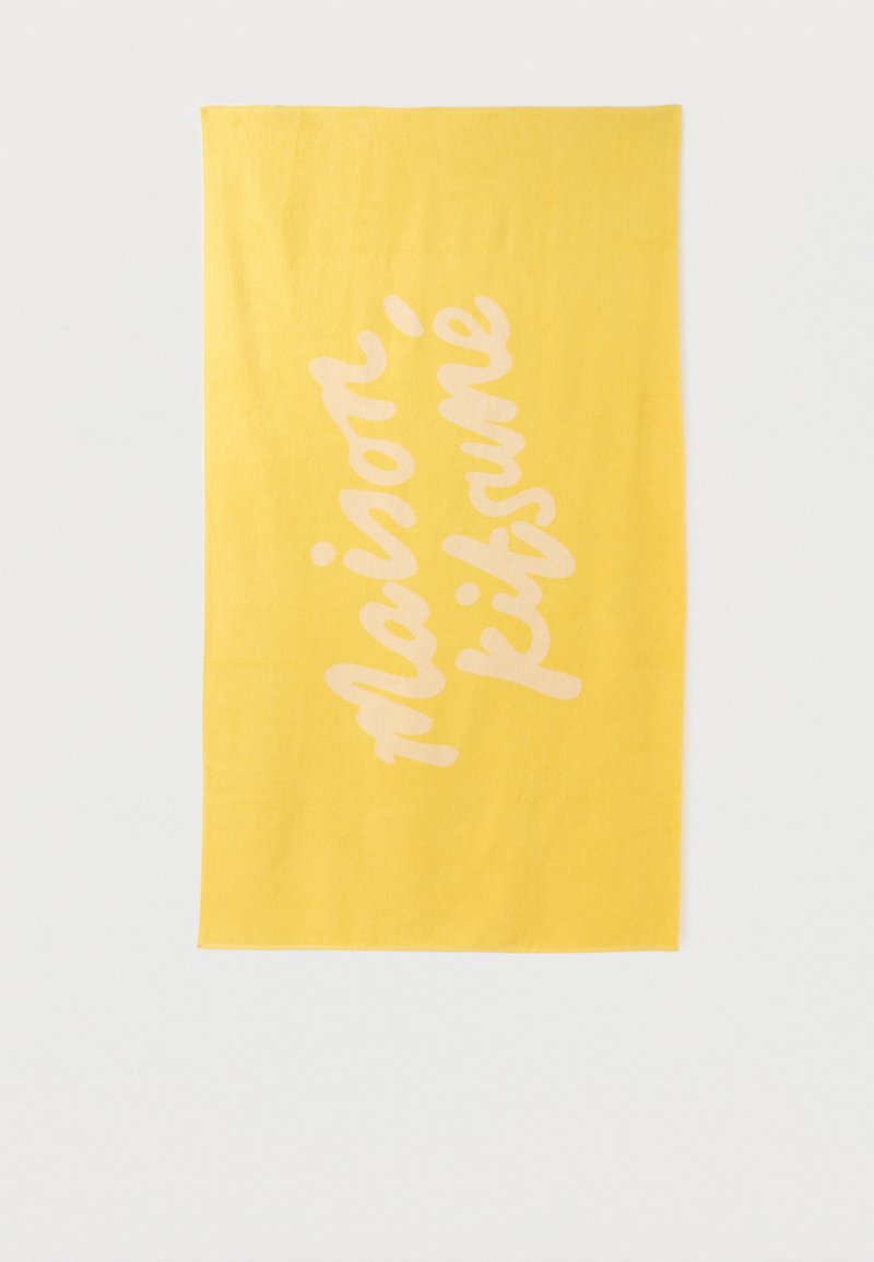 Κίτρινη ορθογώνια πετσέτα με το κείμενο "maison kitsuné" γραμμένο διαγώνια με ανοιχτό ροζ καλλιγραφική γραμματοσειρά.