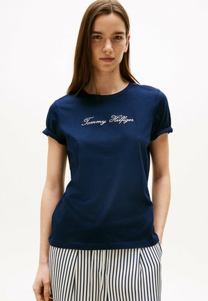 T-shirt bleu marine à manches courtes avec poignets roulés, arborant un logo "Tommy Hilfiger" brodé en blanc sur la poitrine. Tissu en coton doux.