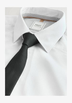 Valge dressipluus tekstuuriga musta lipsuga. Pluusil on sile kangas ja mustrit sisaldav sisekaelus. Sildil on kiri "NEXT TAILORING."