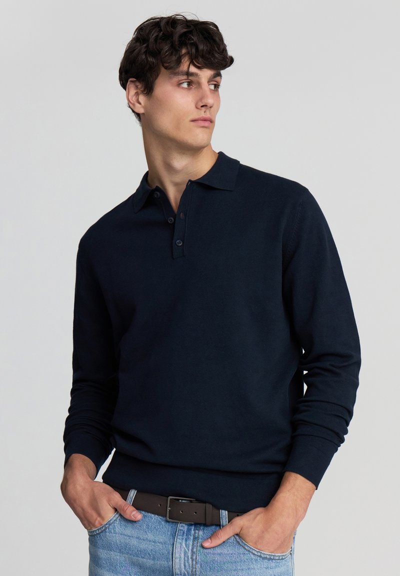Polo a maniche lunghe blu navy con colletto, caratterizzato da dettagli con bottoni. Il tessuto appare liscio e a costine ai polsini.