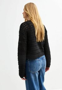 Maglione lavorato a maglia nero con motivo intricato e righe testurizzate, abbinato a jeans a vita alta blu chiaro. Dettagli visibili sulla schiena e lunghezza.