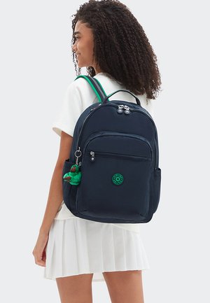 Joven con cabello rizado que lleva un vestido blanco y una mochila azul marino con detalles verdes y un llavero pequeño de mono verde.