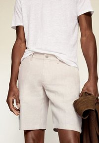 Helle beige Leinen-Shorts mit Fronttaschen, Knopfverschluss und geradem Schnitt, kombiniert mit einem weißen T-Shirt und einer braunen Jacke.