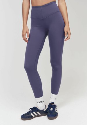 Kobieta ubrana w wysokie purpurowe legginsy, białe skarpetki i granatowe buty sportowe Adidas stoi na jednolitym tle.