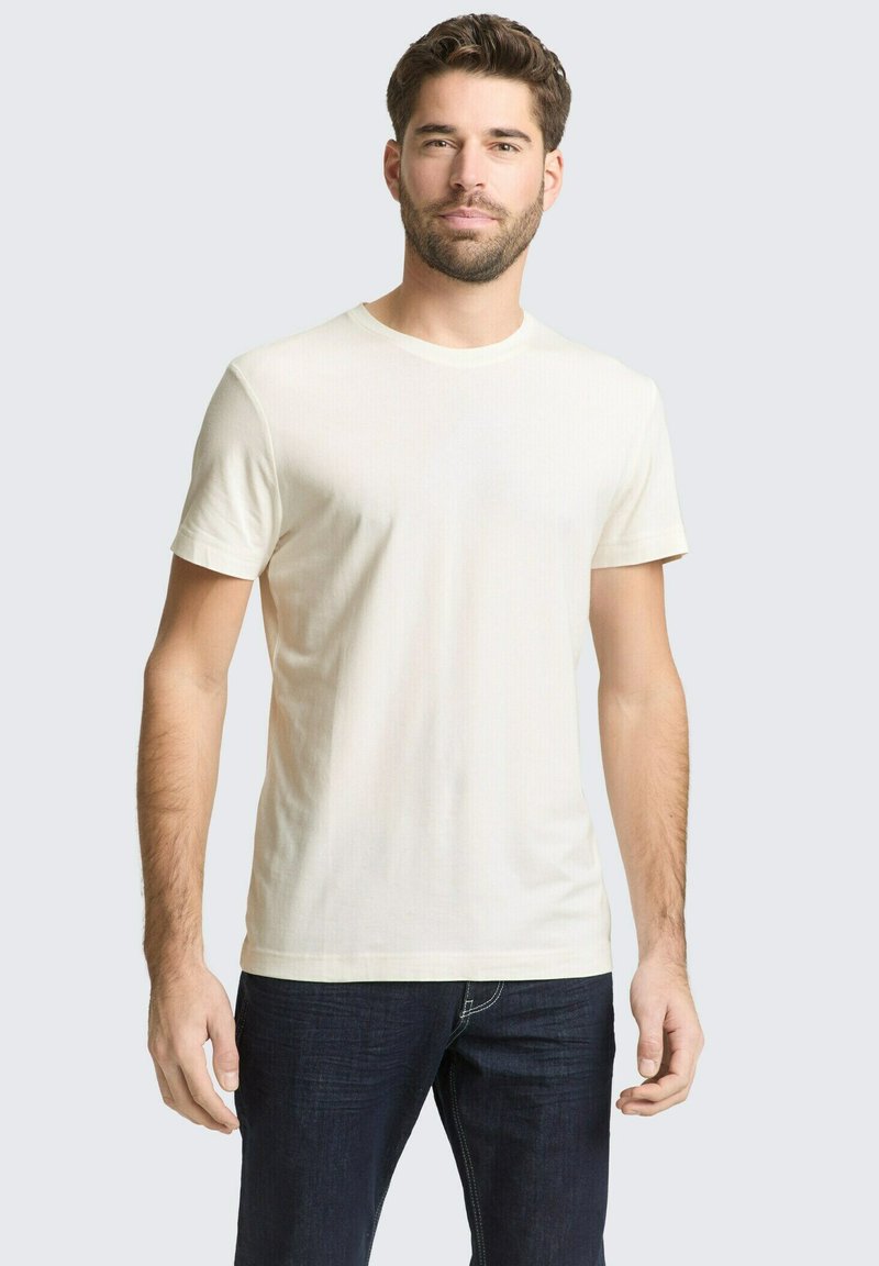 Mann mit kurzem braunem Haar und Bart, trägt ein schlichtes, off-weißes T-Shirt mit kurzen Ärmeln und dunkelblaue Jeans, steht mit entspannten Armen.