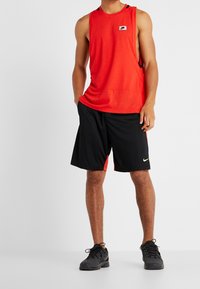 Mann, der ein rotes ärmelloses Nike-Oberteil, schwarze und rote Nike-Shorts sowie schwarze Nike-Sportschuhe trägt und vor einem einfarbigen Hintergrund steht.