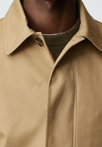 Beige trenchcoat met een spitse kraag, voorzien van een bruine knoop en een zichtbare groene geribbelde trui eronder. Glad oppervlak.