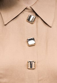 Μπεζ πουκάμισο με γιακά και τρία μεγάλα τετράγωνα μεταλλικά κουμπιά τοποθετημένα κατακόρυφα στο μπροστινό μέρος.
