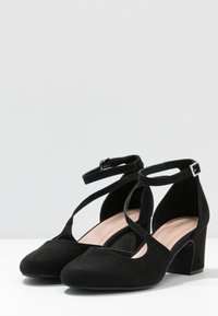 Zapatos de tacón alto de ante negro con una correa en el tobillo y lados abiertos, descansando sobre una superficie blanca contra un fondo neutro.
