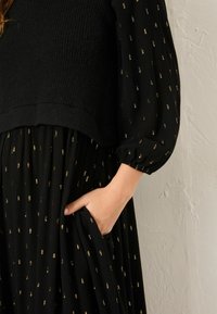 Schwarzes Kleid mit einem strukturierten Strickoberteil und einem fließenden Rock mit goldenen Punktemustern. Lange Ärmel mit elastischen Bündchen und Taschendetails.