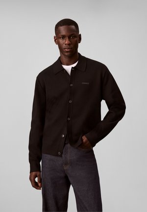 Homme portant un cardigan noir boutonné sur une chemise blanche avec un jean foncé, debout devant un fond gris uni.