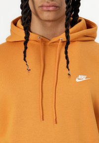 Moletom Nike em laranja brilhante, com um bolso canguru frontal, capuz com cordão e pequeno logo branco no peito. Tecido de textura suave.