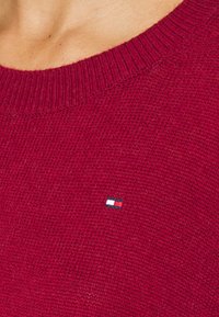 Gros plan sur une personne portant un pull rouge en tricot avec un petit logo rectangulaire blanc, rouge et marine sur la poitrine.