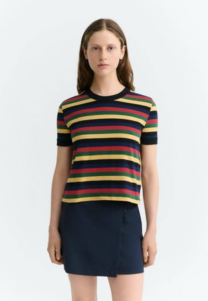 Femme aux longs cheveux portant une chemise à manches courtes avec des rayures horizontales jaunes, rouges, vertes et bleu marine et une jupe bleu marine foncé.