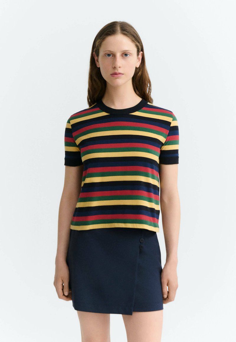 Femme aux longs cheveux portant une chemise à manches courtes avec des rayures horizontales jaunes, rouges, vertes et bleu marine et une jupe bleu marine foncé.
