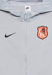 Ανοιχτό γκρι ζακέτα με φερμουάρ, με μαύρο λογότυπο Nike στην αριστερή πλευρά και πορτοκαλί έμβλημα ασπίδας με λιοντάρι και γράμματα "CFC" στη δεξιά πλευρά του στήθους.