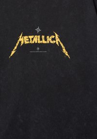 Černé tričko s opotřebovaným žlutým logem "Metallica" s ostrohrannými okraji a prasklinami, tištěné na přední straně uprostřed. Měkká textura.