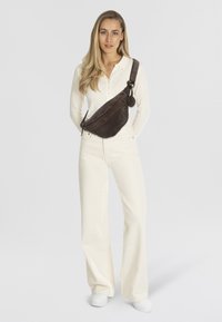 Borsa a tracolla in pelle marrone con design curvato, chiusura lampo, tracolla regolabile e accento in metallo. Il modello indossa una camicia bianca e pantaloni.