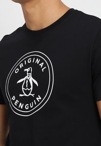 Camiseta negra con un logo circular blanco de Original Penguin que presenta un pingüino estilizado con corbata en el pecho.