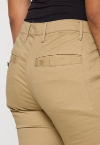 Gros plan sur un pantalon beige ajusté avec des poches passepoilées à l'arrière, porté par une personne en haut noir, montrant les coutures détaillées et la texture du tissu.