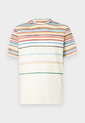 Muž stojaci s rukami v vreckách, oblečený v krémovom tričku s viacfarebnými pruhmi, tmavohnedých nohaviciach, červenom páse a čiernych topánkach.