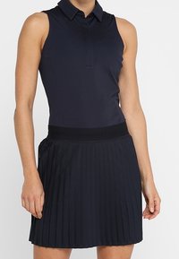 Polo senza maniche in blu navy con colletto, abbinato a una gonna plissettata blu navy. La gonna presenta una vita elastica con logo.