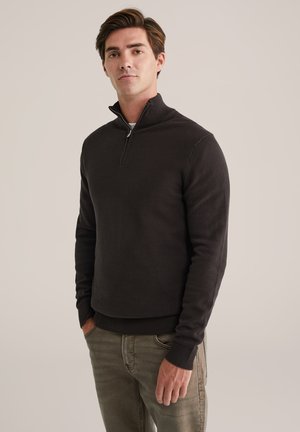 Suéter de cuello alto de color marrón oscuro con tejido texturizado, puños y dobladillo acanalados, combinado con jeans de color verde oliva claro. Diseño simple y elegante.