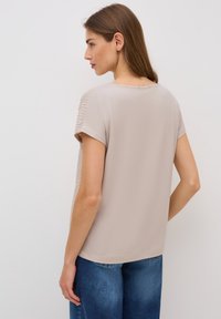 Blusa beige de manga corta con una textura suave y acentos acanalados en los hombros. Conjunto mostrado con jeans de mezclilla azules.