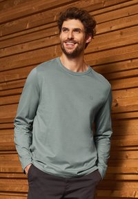 Camisa de manga larga de color verde claro, hecha de una tela suave, con un cuello redondo y un sutil detalle de logo en el pecho.