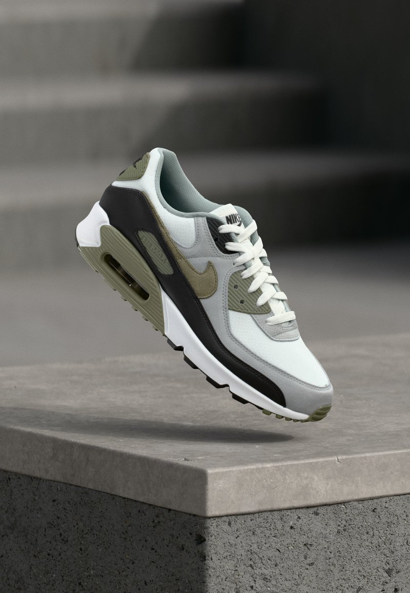 Scarpa Nike Air Max nei colori bianco, nero e verde oliva appoggiata su un bordo di cemento con scalini sfocati sullo sfondo.