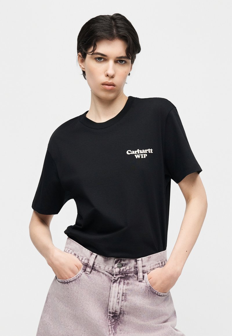 Junge Person mit kurzen dunklen Haaren, die ein schwarzes Carhartt WIP T-Shirt und helllila Jeans mit ausgewaschenem Look trägt, die Hände in den Taschen.