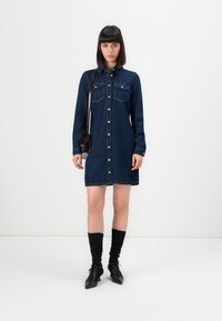Femme debout dans une robe en denim bleu à boutons, avec des chaussettes noires, des chaussures à talons noires et un sac à épaule noir, devant un fond blanc uni.