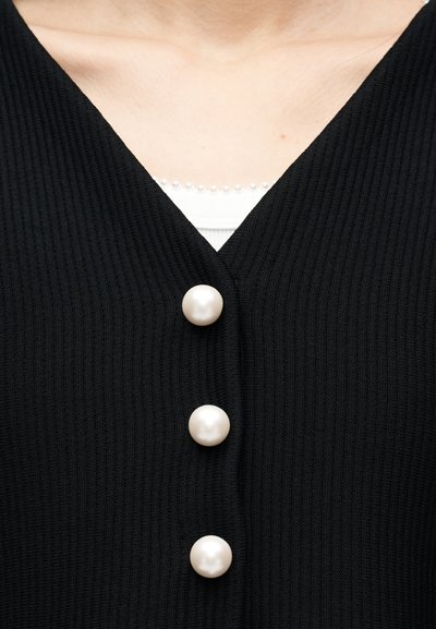 Κοντινό πλάνο μαύρου ριμπ πλεκτού ρούχου με τρία μεγάλα κουμπιά από μαργαριτάρι και άσπρο υποκάμισο με μικρή διακοσμητική λεπτομέρεια από μαργαριτάρια στη λαιμόκοψη.
