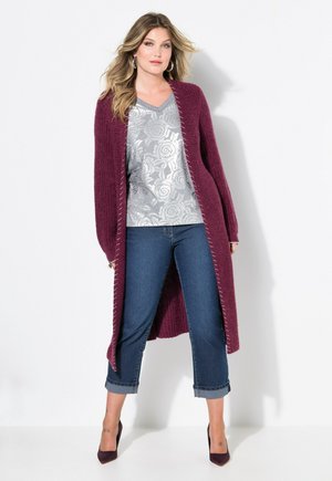Cardigan lungo bordeaux in maglia con apertura davanti, sovrapposto a una blusa con stampa floreale argentata e jeans blu a vita alta. Tacchi neri completano il look.