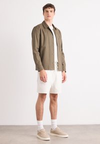 Khaki, napapahid na jacket na may kuwelyo at bulsa sa gilid, suot sa itaas ng puting t-shirt at beige na shorts. Naka-beige na sneakers ang modelo at may puting medyas.