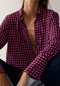 Femme portant une chemise à motifs maroon, marine et blanche à manches longues avec le col ouvert, assise les jambes croisées dans un pantalon noir.