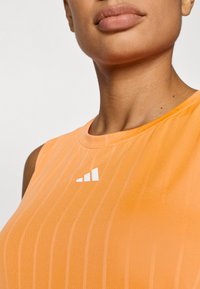 Πορτοκαλί αθλητικό τοπ με πλέγμα υφή, κάθετες ρίγες και λευκό λογότυπο στο στήθος, κοντινή θέα στον λαιμό και στους ώμους.
